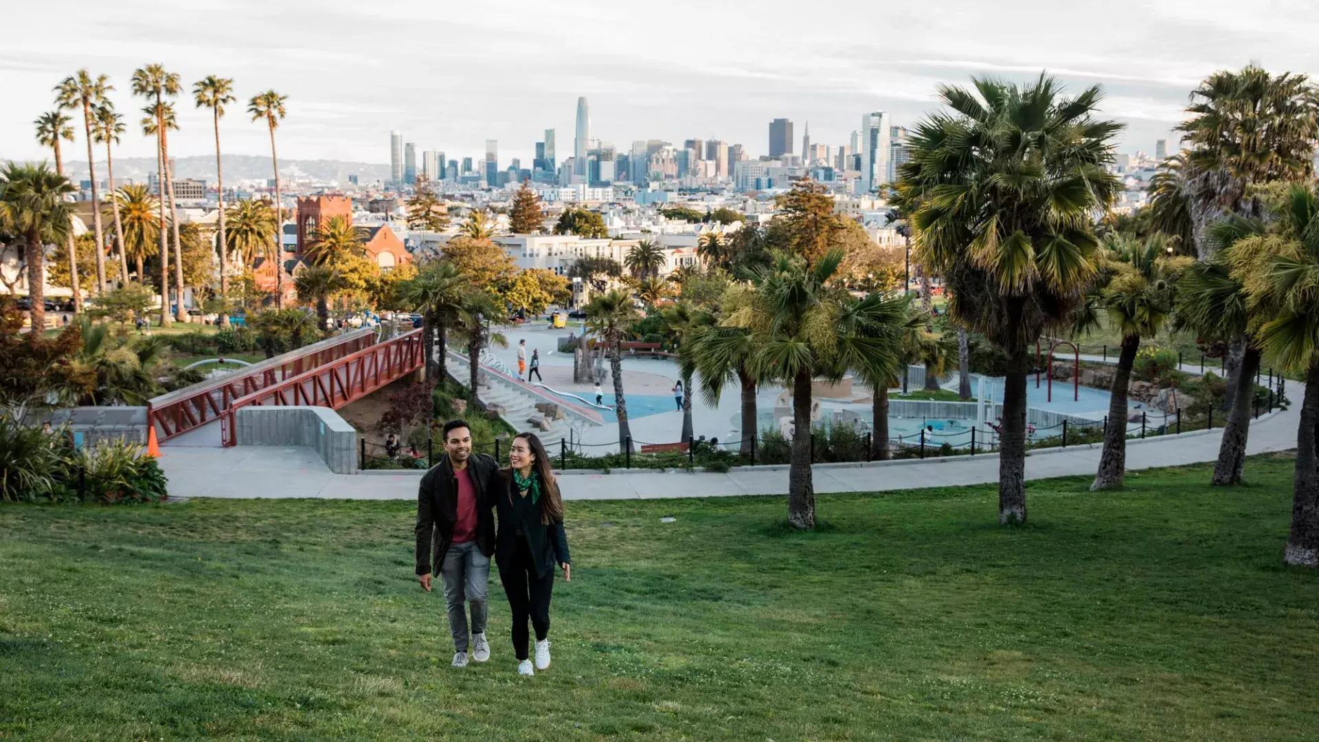 ドロレス公園とサンフランシスコのスカイラインを背にカメラに向かって歩くカップル。