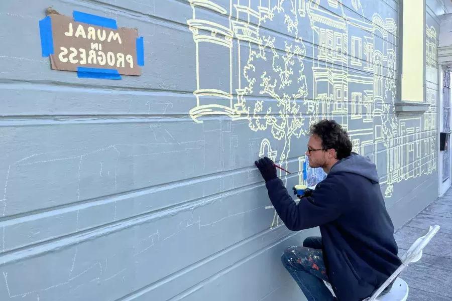 An artist paints a mural on the side of a building in the 任务的区, with a sign taped onto the building that reads "Mural in Progress." San Francisco, CA.
